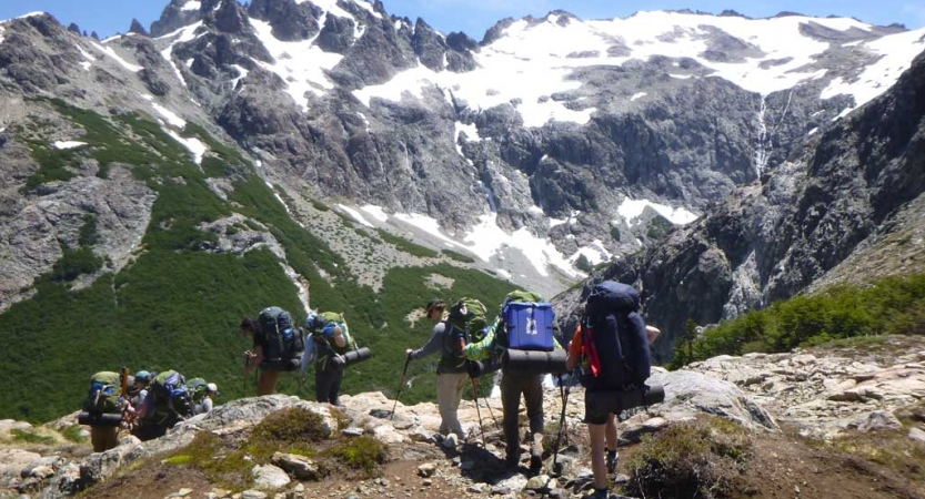 A group of people wearing backpacks hike through a mountainous landscape, with snow-capped mountains in the background. 